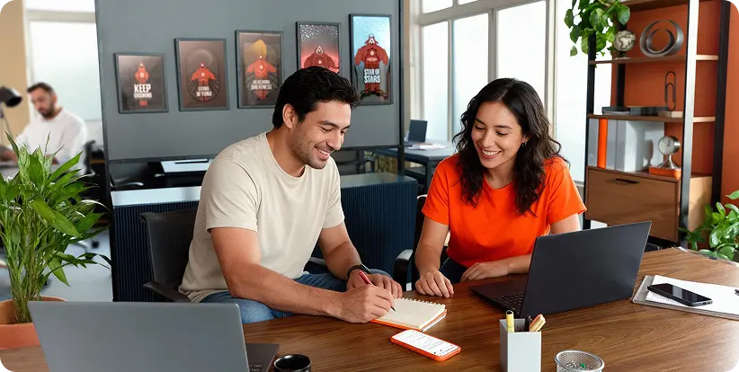 Dos emprendedores sonríen mientras trabajan juntos en una oficina con sus portátiles, representando el acompañamiento de Hotmart para quienes escalan productos digitales.