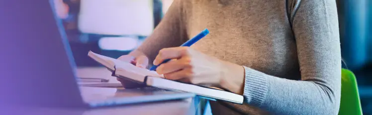 A person wearing a gray sweater taking notes in a notebook while sitting at their computer.