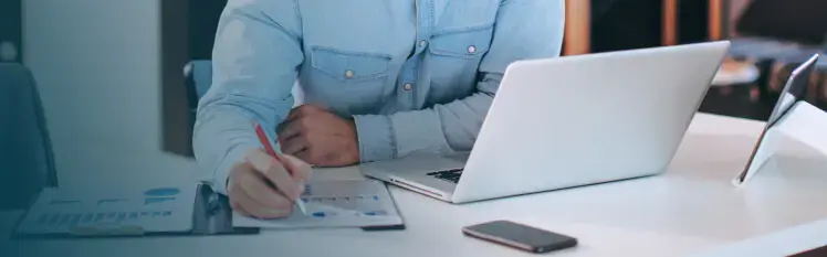 A person in blue, long-sleeved shirt writing in their notebook with a laptop to their left.